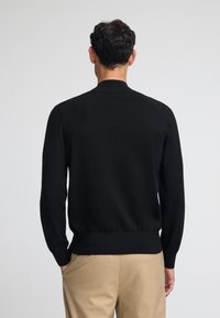 Svart stickad kofta med hög krage, långa ärmar och figursydd midja, kombinerad med beige byxor. Slät struktur, minimalistisk design.