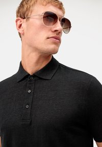 Schwarzes Herren-Polohemd mit klassischem Kragen, Dreiknopfleiste und glatter Textur. Modell trägt getönte Fliegerbrille.