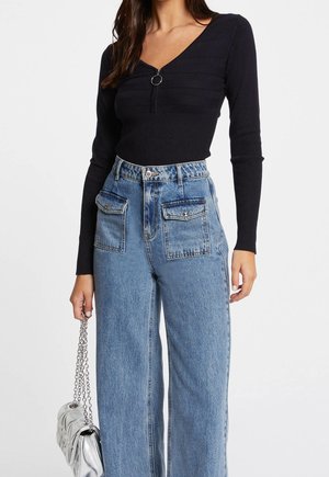 Kvinde iført sort, ribbet langærmet top med ringlynlås og højtaljede blå jeans med store frontlommer, der holder en sølvfarvet håndtaske.