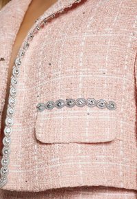 Jachetă roz din tweed cu un model texturat, având o buzunar și embellishments argintii de-a lungul gulerului și marginilor buzunarului.
