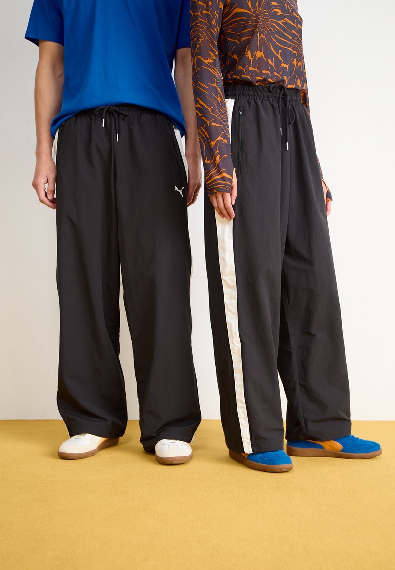 Sorte, løstsittende bukser med elastisk midje, prydet med side-striper, kombinert med fargerike skjorter og varierte sneakers på et gult gulv.