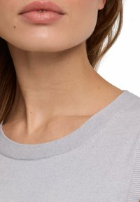 Maglione in maglia grigio chiaro con scollatura ampia. Ripresa ravvicinata che mette in risalto la morbida texture e i toni naturali della pelle. Nessun motivo o accessorio visibile.