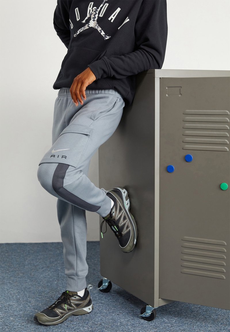 Grå joggers med cargo-fickor och mörka sidpaneler, matchade med svarta och gröna sportskor. Svart sweatshirt med grafisk design.