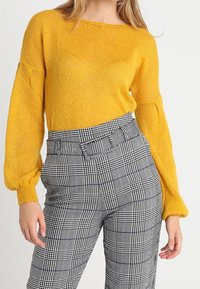 Gul stickad tröja med vid ringning och puffärmar, kombinerad med svart och vit rutmönstrad högmidjad byxa med bälte.
