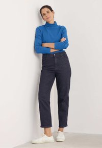 Blauwe geribde turtleneck, donkere jeans en witte sneakers. De top heeft lange mouwen; de jeans zijn recht gesneden met een hoge taille.