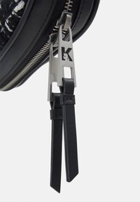 Tirette de fermeture éclair en cuir noir avec une étiquette en métal portant la lettre "K", accompagnée de deux sangles en cuir noir.