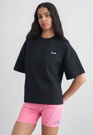 Hosszú sötét hajú nő fekete ellesse pólót és rózsaszín ellesse kerékpáros rövidnadrágot visel, világos egyszínű háttér előtt áll.