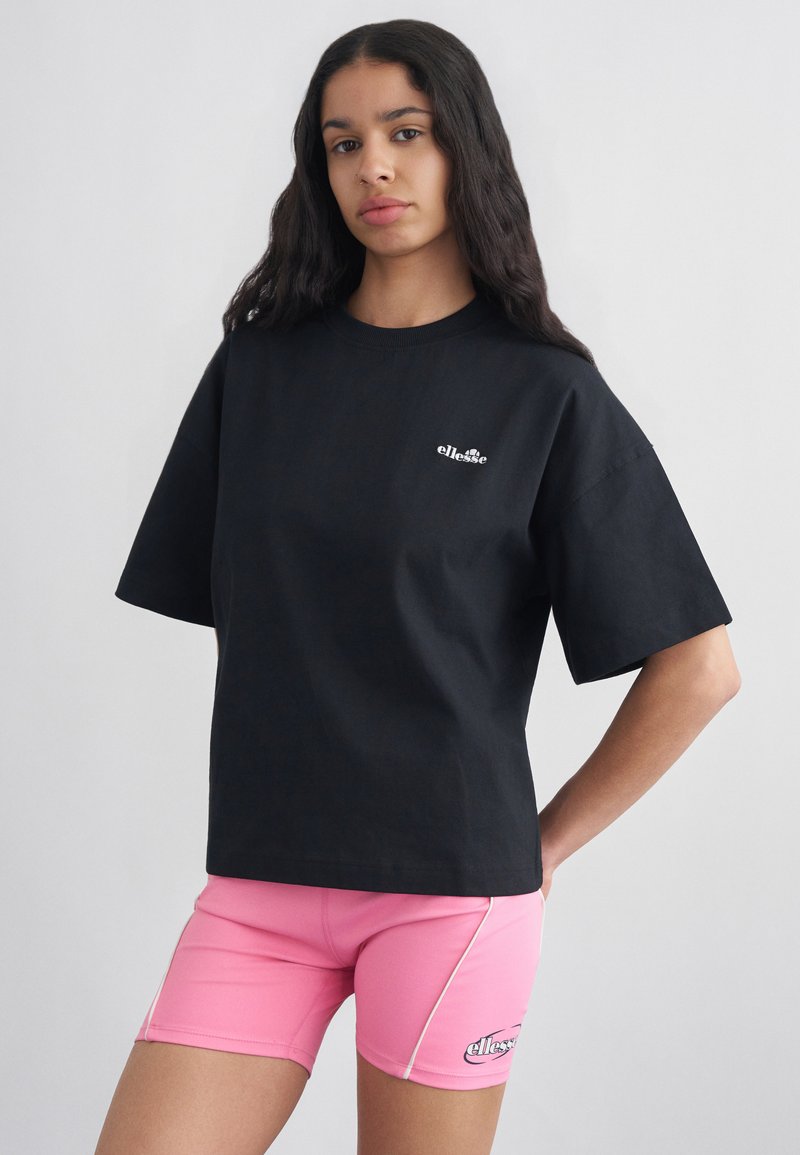 Γυναίκα με μακριά σκούρα μαλλιά φοράει μαύρο t-shirt ellesse και ροζ ποδηλατικά σορτς ellesse, στέκεται μπροστά σε απλό φωτεινό φόντο.