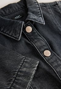 Veste en jean noire avec un col pointu, deux boutons marron et des surpiqûres contrastantes. Le matériau présente une texture légèrement usée.