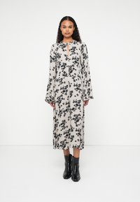 Robe midi à fleurs en beige avec un motif floral noir, manches longues, décolleté en trou de serrure et une silhouette droite. Associée à des bottines noires.