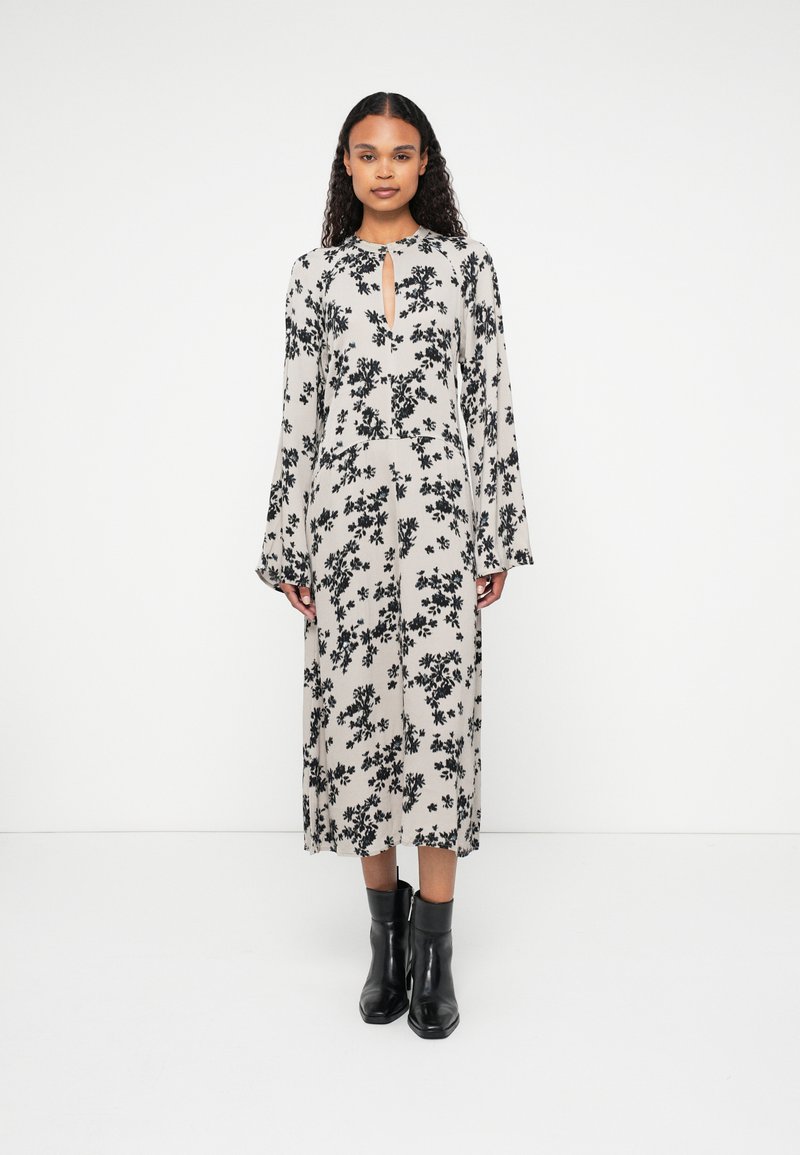 Robe midi à fleurs en beige avec un motif floral noir, manches longues, décolleté en trou de serrure et une silhouette droite. Associée à des bottines noires.