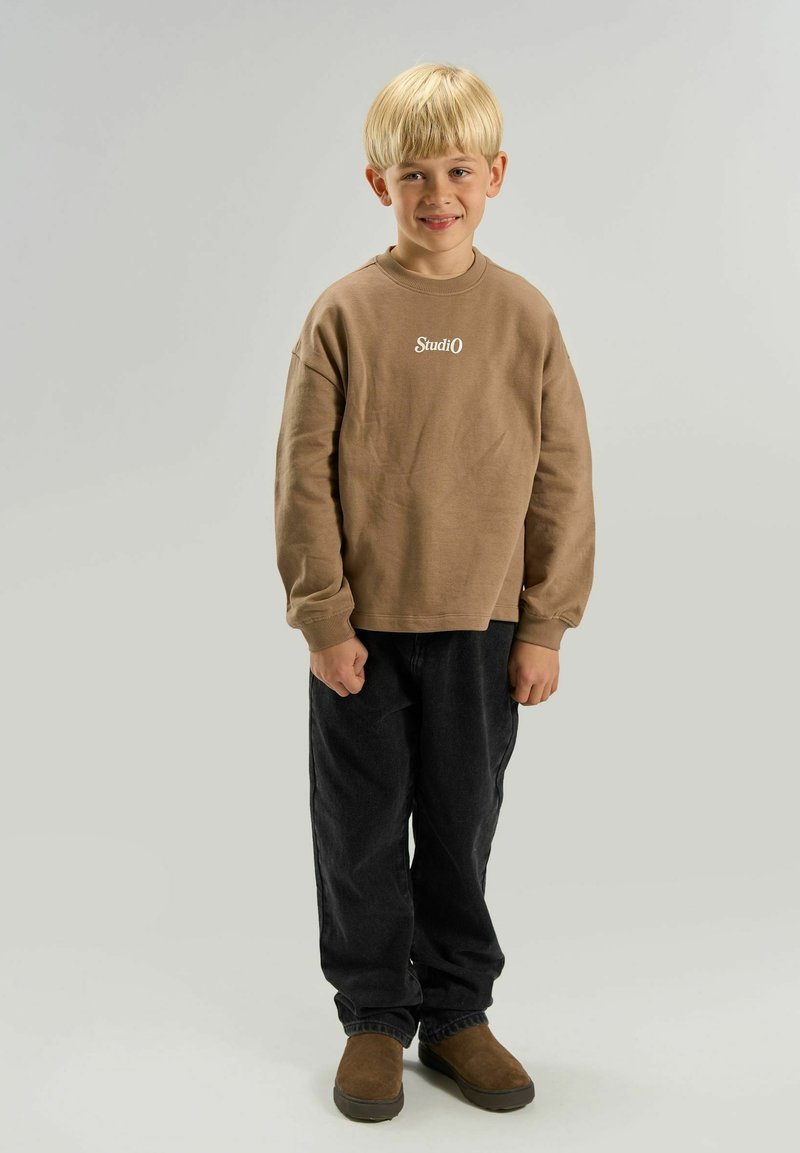 Garçon blond debout et souriant, portant un t-shirt marron à manches longues avec le texte « StudiO », pantalon noir et chaussures marron sur un fond gris uni.