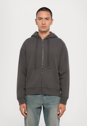 Sweatshirt cinzento com fecho, bolso tipo canguru, capuz ajustável com cordão e punhos canelados, combinado com jeans azul claro.