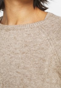 Pull en maille beige avec un col rond côtelé, présentant une texture douce et de légères variations de couleur sur l'ensemble du tissu.