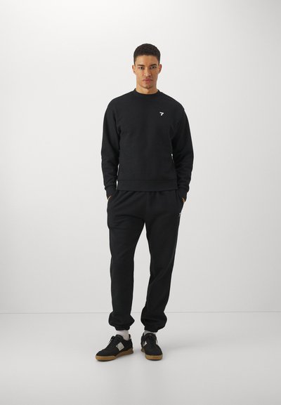 Sudadera negra y pantalones de chándal, mezcla de algodón, con puños acanalados y un pequeño logo blanco en el pecho. Los zapatos son negros con suelas de goma.
