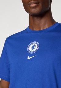 Blå bomulds t-shirt med rund hals. Har et Chelsea FC logo og Nike swoosh i hvidt, med korte ærmer og en glat tekstur.