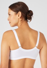 Sosten blanco de tela suave, con bordes festoneados, tirantes ajustables y un diseño liso. Presenta elementos metálicos para el ajuste de los tirantes.