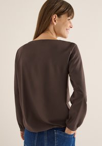 Blusa marrone a maniche lunghe con vestibilità ampia, scollatura rotonda e polsini elasticizzati. Texture liscia, design minimalista, abbinata a jeans di denim blu.