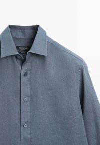 Blauw linnen overhemd met lange mouwen en knopen, met kraag, gedeeltelijk dichtgeknoopt, hangend tegen een witte achtergrond.