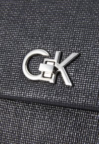 Černá taška z texturované kůže s lesklou stříbrnou sponou se znaky "CK", která nabízí moderní design.