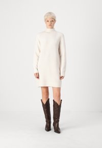 Πλεκτό φόρεμα σε off-white, με ψηλό γιακά και μακριά μανίκια. Συνοδευόμενο από καφέ δερμάτινες καουμπόικες μπότες με διακοσμητική ραφή.