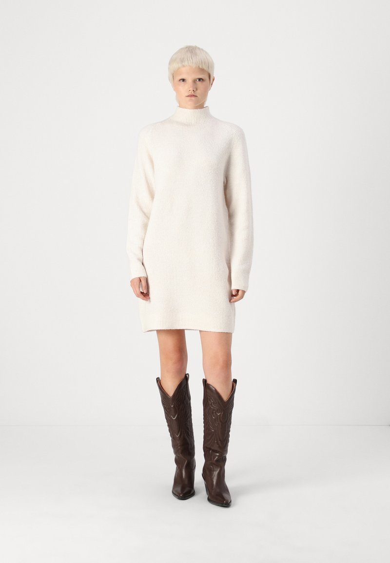 Πλεκτό φόρεμα σε off-white, με ψηλό γιακά και μακριά μανίκια. Συνοδευόμενο από καφέ δερμάτινες καουμπόικες μπότες με διακοσμητική ραφή.