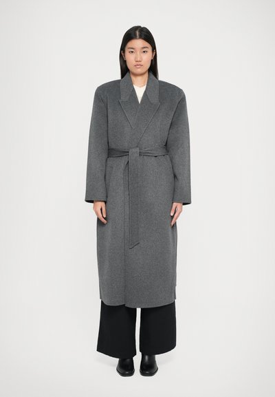 Grauer Wollmantel mit langen Ärmeln, Reverskragen und Gürtel in der Taille. In einer oversized Form gestaltet, kombiniert mit schwarzen weit geschnittenen Hosen und Stiefeln.