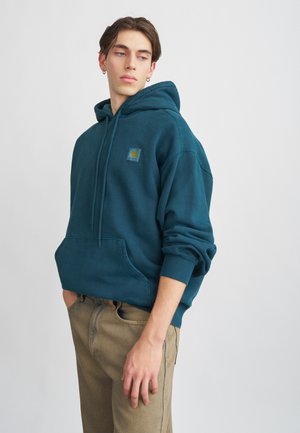 Türkisfarbenes Baumwoll-Hoodie mit einer Fronttasche, gerippten Bündchen und einem kleinen Logopatch auf der linken Brust. Trägt dazu hellbraune Hosen.