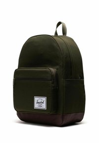 Sac à dos en tissu vert foncé de forme arrondie. Compartiment principal unique, poche avant zippée et base en cuir marron. Doté d'un écusson logo.