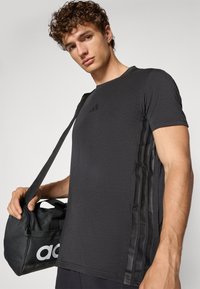 Schwarzes Sport-T-Shirt mit kurzen Ärmeln, das über strukturierte Einsätze an den Seiten und ein dezentes Logo auf der Brust verfügt. Trägt eine schwarze Tasche.