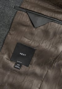 Doublure de costume marron à motif avec étiquette en tissu noire indiquant "NEXT," "SLIM FIT," taille "40R" et "MADE IN BANGLADESH."