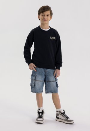 Sweat à capuche noir avec un texte graphique, associé à un short cargo en denim bleu et des sneakers noires, blanches et grises. Se tenant devant un fond uni.
