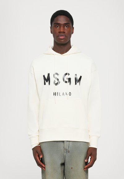 Jeune homme portant un sweat à capuche MSGM Milano blanc cassé et un jean vert délavé, se tenant devant un fond blanc uni.