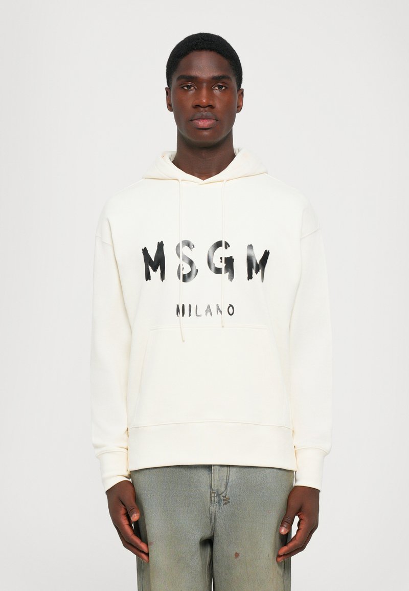 Giovane uomo che indossa una felpa MSGM Milano bianco sporco e jeans verdi sbiaditi, in piedi davanti a uno sfondo bianco semplice.