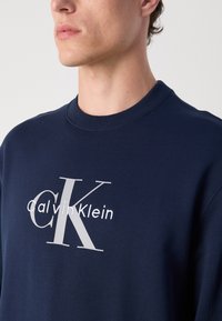 Sweatshirt azul-marinho feito de algodão, com um decote redondo e um grande logotipo da Calvin Klein impresso em cinza na parte da frente.