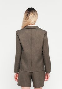 Žena s dlhými blond vlasmi, oblečená v hnedej prispôsobenej saku a zodpovedajúcich šortkách, otočená chrbtom proti jednému bielému pozadiu.