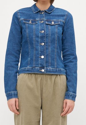 Personne portant une veste en jean bleue avec des boutons argentés et un pantalon en velours beige, vue du cou aux hanches sur un fond uni.