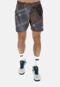 Jambes masculines portant un short à motifs géométriques multicolores, des chaussettes blanches et un avis de sport bleu marine et blanc, se tenant devant un fond uni.