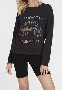Musta puuvillapaita, jossa on värikäs graafinen kuvasto, jossa on kaksi kasvot, joissa on avaruusoliomaista ilmettä ja salamoita. Teksti lukee "ALIENATED ALIEN YOUTH."