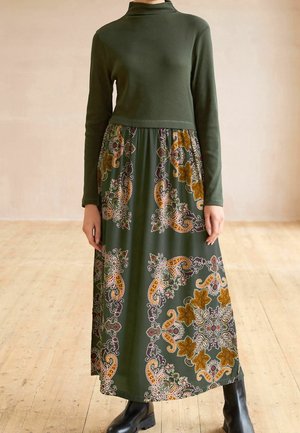 Robe verte à manches longues avec un haut ajusté et une jupe fluide à motif paisley en or, violet et vert. Bottes noires.