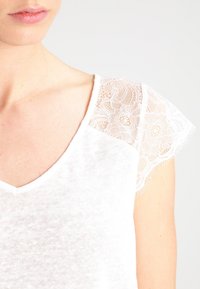 Bílý tričko s měkkým, texturovaným tělem a krajkovými krátkými rukávy s vlnitým lemem, které zobrazuje design s výstřihem do V.