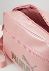 Borsa sintetica rosa con una tasca anteriore con zip, texture liscia e logo Puma in argento metallizzato. La zip aperta rivela un interno foderato.