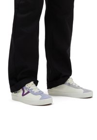 Sneakers Vans con toma in crema, dettagli in suede lavanda e suola in gomma bianca, abbinati a pantaloni neri con orlo arrotolato.