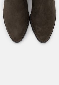 Botines de ante marrón con diseño de punta afilada, que presentan una textura suave y costuras sutiles a lo largo de los bordes.