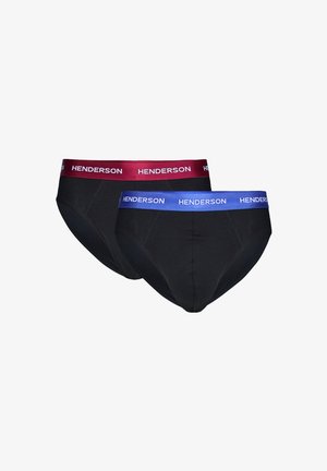 Oberkörperfreier Mann schaut nach rechts, richtet sein Handgelenk, trägt schwarze Slips mit bordeauxrotem Bund, auf dem "HENDERSON" steht, vor neutralem Hintergrund.