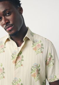 Camicia a maniche corte color crema con righe verticali e ricami floreali in rosa, blu e verde. Colletto con bottoni e tessuto texturizzato.