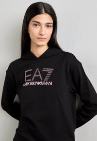 Fekete kapucnis pulóver, amelyen strasszkövekből készült "EA7" logó és rózsaszín írásban "EMPORIO ARMANI" felirat található, puha anyagból készült és laza szabású.