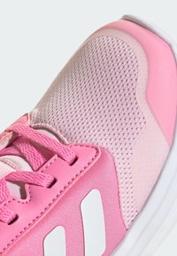 Różowy but sportowy z siateczki i materiałów syntetycznych, teksturowana cholewka, różowe sznurówki i białe akcenty w paski po bokach.