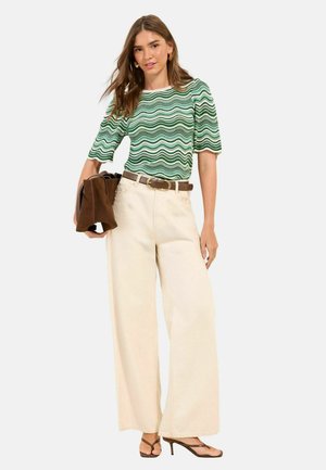 Žena v zeleno-bielom tričku s vlnkovým vzorom, širokých krémových nohaviciach, hnedom opasku, sandáloch a s hnedou taškou v ruke.