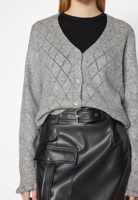 Szary dzianinowy kardigan z diamentowym wzorem, dekoltem w serek i jasnoróżowymi guzikami, zestawiony z czarną skórzaną spódnicą z kieszenią naszywana.